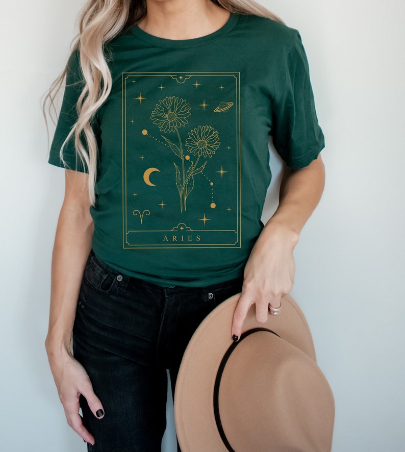 May include: A green t-shirt with a gold graphic of a flower with a crescent moon and stars. The graphic is in a rectangular frame with the text "ARIES" at the bottom.