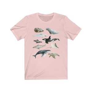 K&ouml;nnte beinhalten: Ein rosa T-Shirt mit einer Aquarell-Illustration verschiedener Meerestiere, darunter ein Wal, ein Delfin, eine Meeresschildkr&ouml;te, ein Seekuh, ein Mantarochen und eine Robbe.