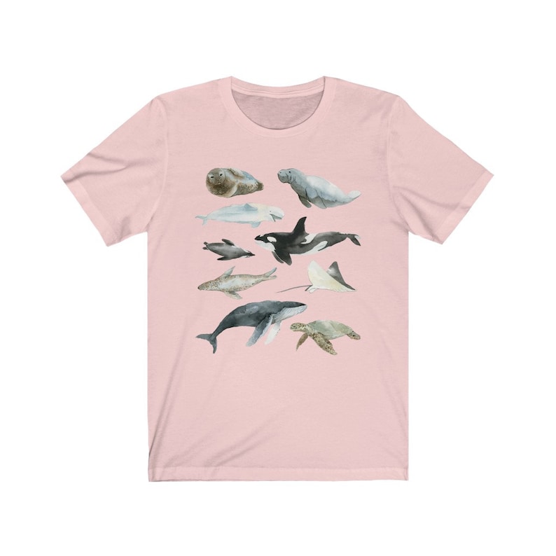 K&ouml;nnte beinhalten: Ein rosa T-Shirt mit einer Aquarell-Illustration verschiedener Meerestiere, darunter ein Wal, ein Delfin, eine Meeresschildkr&ouml;te, ein Seekuh, ein Mantarochen und eine Robbe.