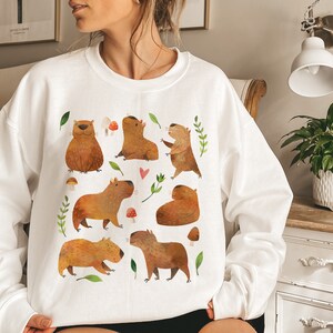 May include: White sweatshirt with a graphic print of multiple capybaras in various poses, surrounded by green leaves and red mushrooms.