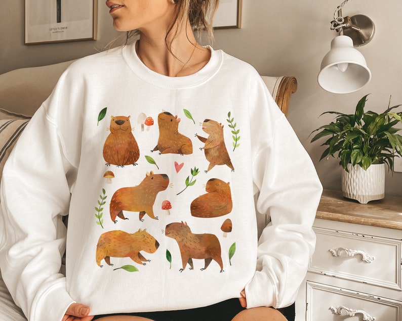 May include: White sweatshirt with a graphic print of multiple capybaras in various poses, surrounded by green leaves and red mushrooms.