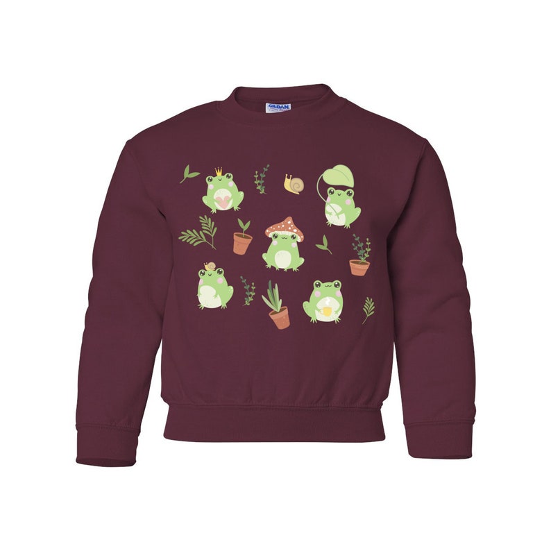 K&ouml;nnte beinhalten: Ein maroonfarbener Pullover mit einem Muster aus gr&uuml;nen Fr&ouml;schen, Pilzen und Pflanzen. Die Fr&ouml;sche tragen Kronen, H&uuml;te und halten Blumen.