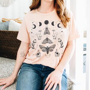 May include: A light pink t-shirt with a black and white graphic print of moths, mushrooms, crystals, and moons. The graphic is centered on the shirt.