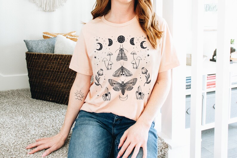 May include: A light pink t-shirt with a black and white graphic print of moths, mushrooms, crystals, and moons. The graphic is centered on the shirt.