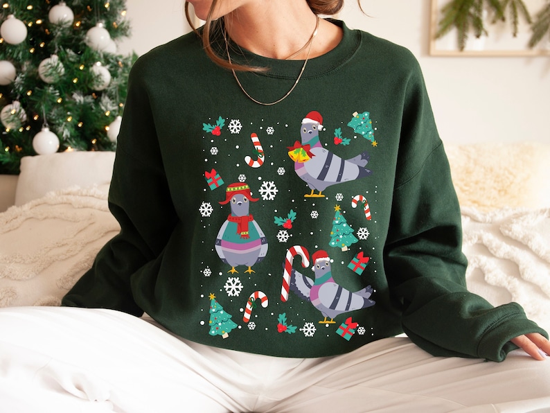 May include: A green Christmas jumper with a festive design featuring cartoon pigeons wearing Santa hats and holding gifts. The jumper has a crew neck and long sleeves.