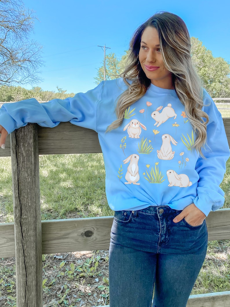 May include: A light blue sweatshirt with a graphic print of white bunnies and flowers. The sweatshirt is worn by a person with long blonde hair.