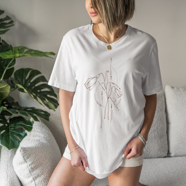 May include: White t-shirt with a brown line drawing of stylized flowers and geometric shapes.