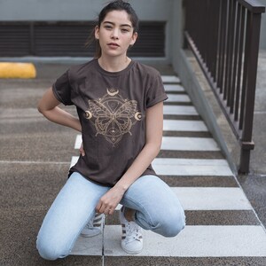 May include: A woman wearing a brown t-shirt with a gold butterfly graphic and blue jeans. The butterfly graphic features a geometric design with moons and stars.
