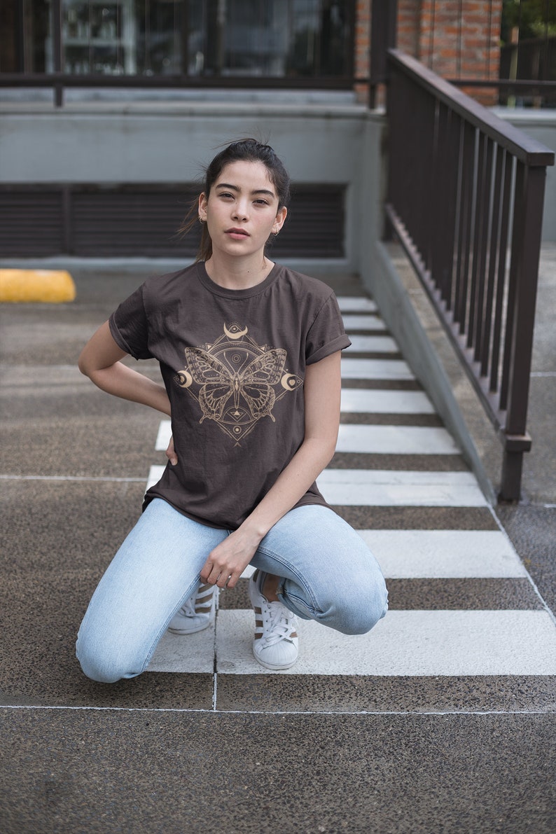 May include: A woman wearing a brown t-shirt with a gold butterfly graphic and blue jeans. The butterfly graphic features a geometric design with moons and stars.