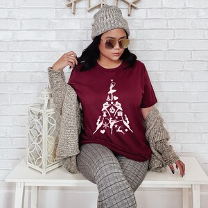 May include: A maroon t-shirt with a white design of a Christmas tree made up of dancers. The tree is decorated with ornaments, stars, and a gift box. The person wearing the shirt is also wearing a grey knitted beanie and sunglasses.