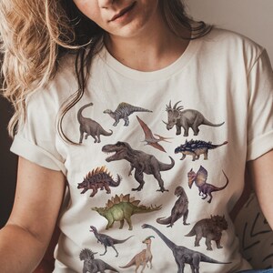 May include: A white t-shirt with a colorful dinosaur print. The dinosaurs are in various poses and sizes, including a Tyrannosaurus Rex, a Triceratops, and a Stegosaurus.