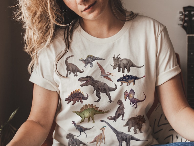May include: A white t-shirt with a colorful dinosaur print. The dinosaurs are in various poses and sizes, including a Tyrannosaurus Rex, a Triceratops, and a Stegosaurus.