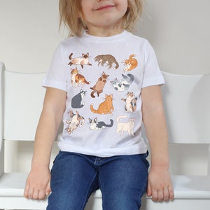 May include: A white t-shirt with a colorful pattern of cats. The cats are in various poses, including sitting, standing, and lying down. The shirt is made of a soft, comfortable material and is perfect for kids.