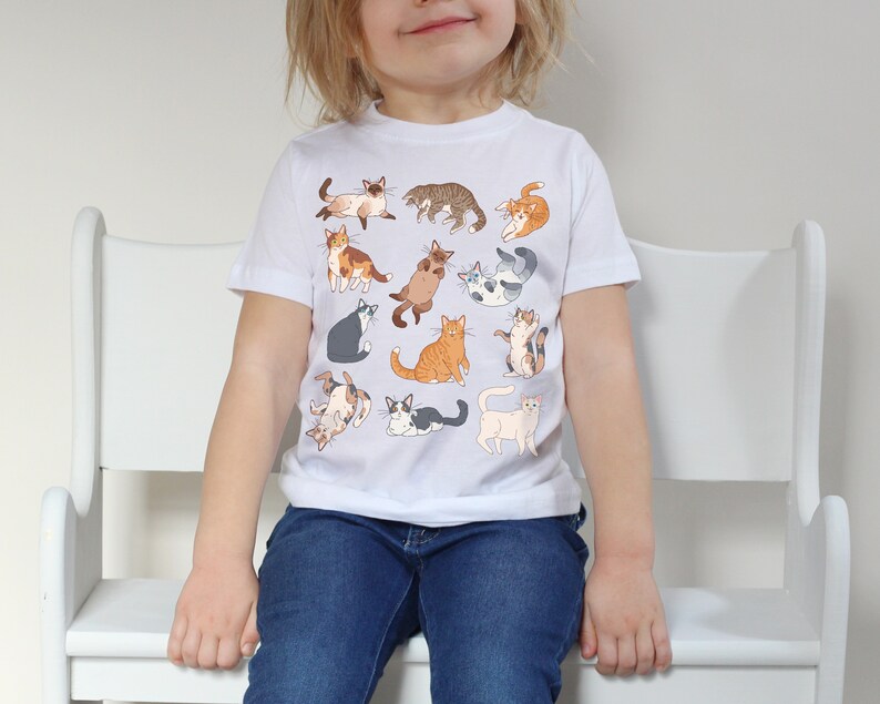 May include: A white t-shirt with a colorful pattern of cats. The cats are in various poses, including sitting, standing, and lying down. The shirt is made of a soft, comfortable material and is perfect for kids.