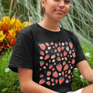 K&ouml;nnte beinhalten: Ein schwarzes T-Shirt mit einem Muster aus roten Erdbeeren und wei&szlig;en Bl&uuml;ten. Das Shirt hat einen Rundhalsausschnitt und kurze &Auml;rmel.