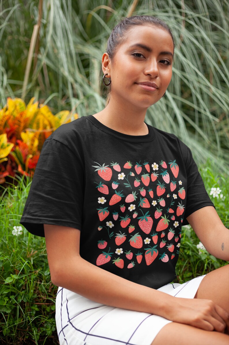 K&ouml;nnte beinhalten: Ein schwarzes T-Shirt mit einem Muster aus roten Erdbeeren und wei&szlig;en Bl&uuml;ten. Das Shirt hat einen Rundhalsausschnitt und kurze &Auml;rmel.