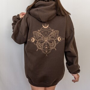 May include: A brown hooded sweatshirt with a graphic print of a butterfly inside a geometric design. The butterfly is outlined in a light brown color and has a crescent moon on each side.