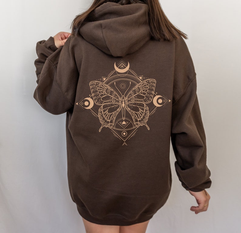 May include: A brown hooded sweatshirt with a graphic print of a butterfly inside a geometric design. The butterfly is outlined in a light brown color and has a crescent moon on each side.