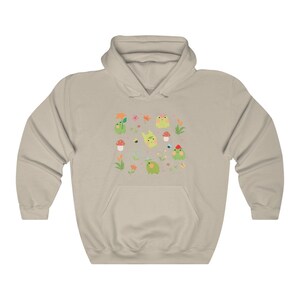 May include: A light beige hoodie with a print of green frogs, flowers, and mushrooms. The print is a repeating pattern and covers the entire front of the hoodie.