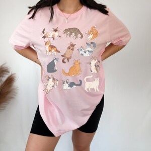 May include: A light pink t-shirt featuring a collection of illustrated cats in various poses. The cats are depicted in a variety of colors and patterns, including tabby, calico, and Siamese. The shirt is a casual, comfortable style.