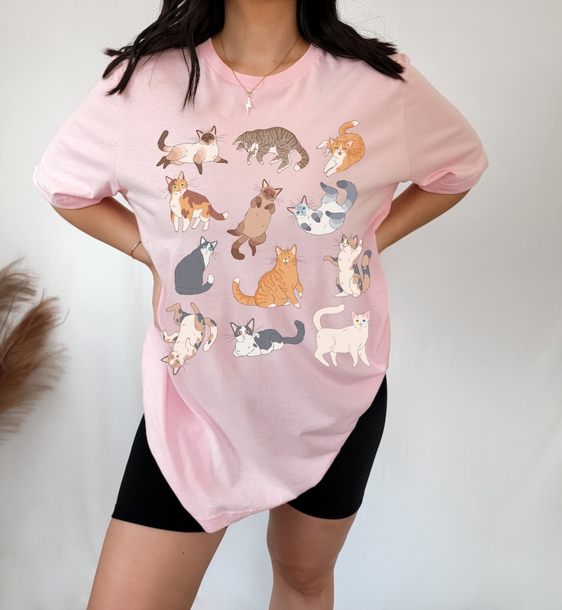 May include: A light pink t-shirt featuring a collection of illustrated cats in various poses. The cats are depicted in a variety of colors and patterns, including tabby, calico, and Siamese. The shirt is a casual, comfortable style.