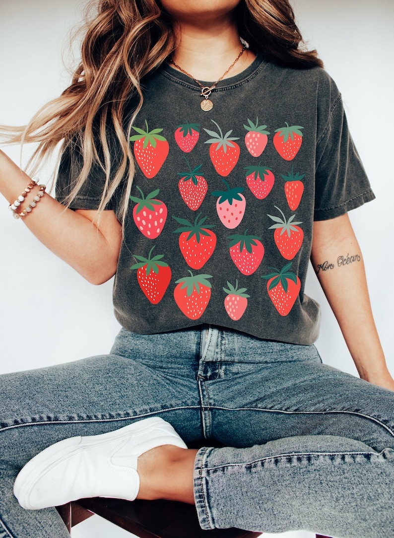 Puede incluir: Una camiseta gris oscura con un estampado de fresas rojas. La camiseta tiene cuello redondo y mangas cortas. La modelo lleva la camiseta con vaqueros azules y zapatillas blancas.