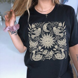 May include: Black t-shirt with a beige floral design featuring birds, butterflies, a sun, and a crescent moon.