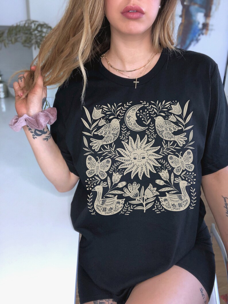 May include: Black t-shirt with a beige floral design featuring birds, butterflies, a sun, and a crescent moon.