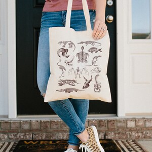 Peut inclure: Un sac fourre-tout beige avec un motif de squelettes d'animaux divers en encre brune. Le sac est tenu par une personne portant un jean bleu et un chemisier rose.