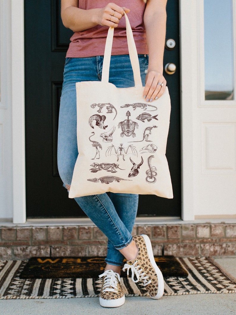 Peut inclure: Un sac fourre-tout beige avec un motif de squelettes d'animaux divers en encre brune. Le sac est tenu par une personne portant un jean bleu et un chemisier rose.