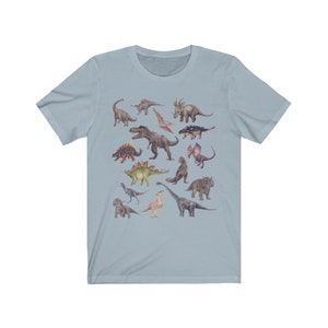 May include: A light blue t-shirt with a colorful illustration of various dinosaurs. The dinosaurs are depicted in a watercolor style and include a variety of species, such as a Tyrannosaurus Rex, a Triceratops, and a Stegosaurus.