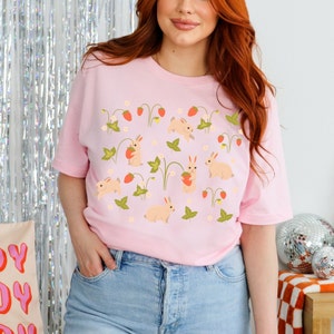 Op de afbeelding: Een roze T-shirt met een patroon van witte konijnen die aardbeien en groene bladeren vasthouden. De konijnen worden omringd door kleine witte bloemen.