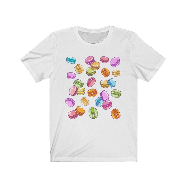May include: White t-shirt with a colorful pattern of macarons. The macarons are in various colors, including pink, blue, green, yellow, and orange.