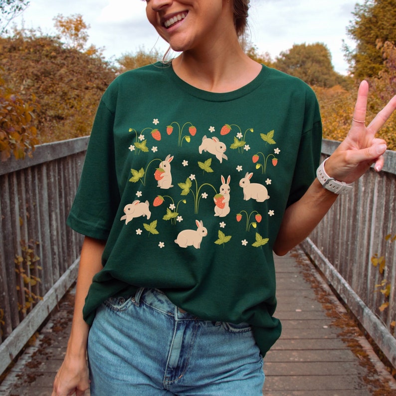 Op de afbeelding: Een donkergroen T-shirt met een patroon van witte konijnen en aardbeien. De konijnen houden aardbeien vast en zijn omringd door groene bladeren en witte bloemen.