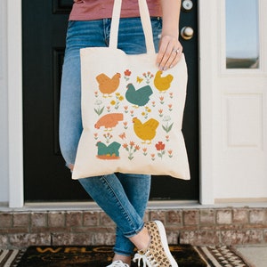 May include: A beige tote bag with a colorful pattern of chickens and flowers. The bag has two long handles and is made of canvas.