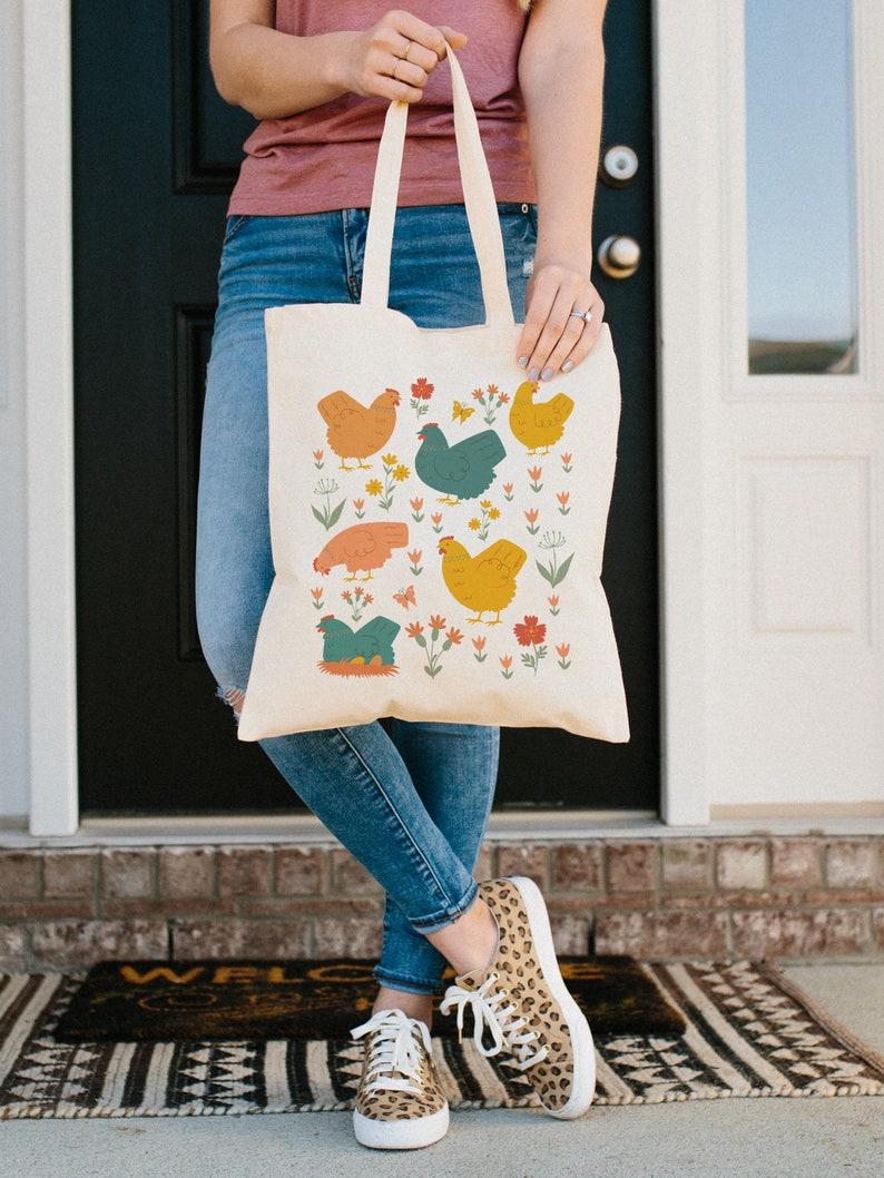 May include: A beige tote bag with a colorful pattern of chickens and flowers. The bag has two long handles and is made of canvas.
