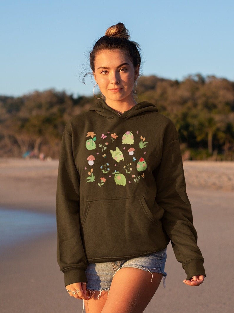 May include: A green hoodie with a pocket on the front. The hoodie has a print of frogs, flowers, and mushrooms in pastel colors.