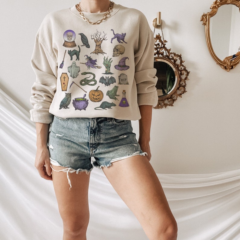 May include: A beige sweatshirt with a Halloween-themed print featuring various spooky icons like a skull, a witch's hat, a cauldron, a ghost, a bat, a snake, a pumpkin, a raven, a cat, and a spider.