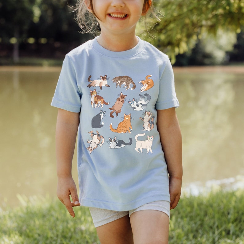 May include: A light blue t-shirt with a pattern of 12 colorful cats in various poses. The shirt is made of a soft, comfortable material.