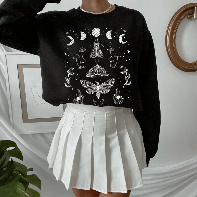 May include: A black cropped sweatshirt with a white graphic print of moths, mushrooms, crystals, and moons. The sweatshirt is paired with a white pleated skirt.