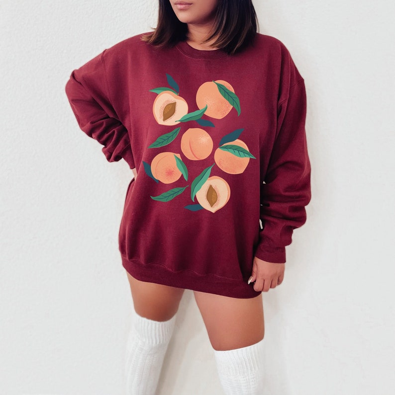 K&ouml;nnte beinhalten: Ein maroonfarbener Pullover mit einem Grafikdruck von Pfirsichen. Die Pfirsiche sind in einer Traube angeordnet und haben gr&uuml;ne Bl&auml;tter.