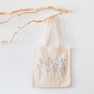 May include: A beige tote bag with a design of blue line drawings of flowers on the front. The bag is hanging on a white wall with a branch of driftwood behind it.