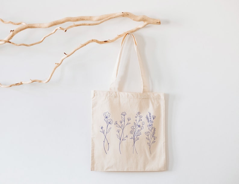 May include: A beige tote bag with a design of blue line drawings of flowers on the front. The bag is hanging on a white wall with a branch of driftwood behind it.