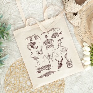 Peut inclure: Un sac fourre-tout beige avec des illustrations brunes de squelettes d'animaux. Les squelettes comprennent un poisson, une tortue, une chauve-souris, un serpent, un oiseau, et plus encore.