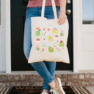 May include: A white canvas tote bag with a colorful design of cartoon frogs, flowers, and mushrooms. The bag has two handles and is being held by a person wearing blue jeans.