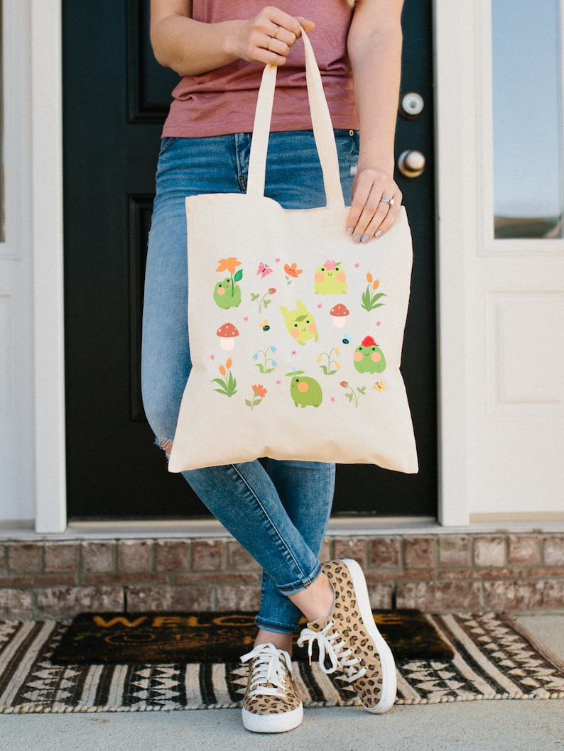 May include: A white canvas tote bag with a colorful design of cartoon frogs, flowers, and mushrooms. The bag has two handles and is being held by a person wearing blue jeans.