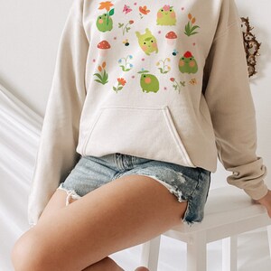May include: A cream-colored hoodie with a graphic print of green frogs, flowers, and butterflies. The hoodie has a drawstring hood and a kangaroo pocket.