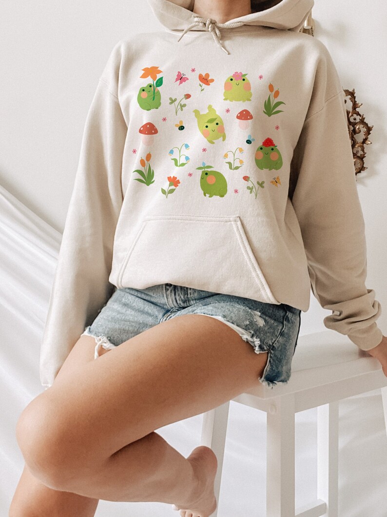 May include: A cream-colored hoodie with a graphic print of green frogs, flowers, and butterflies. The hoodie has a drawstring hood and a kangaroo pocket.