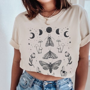 May include: A cream-colored t-shirt with a black and white graphic print of moths, mushrooms, crystals, and crescent moons. The graphic is centered on the shirt.
