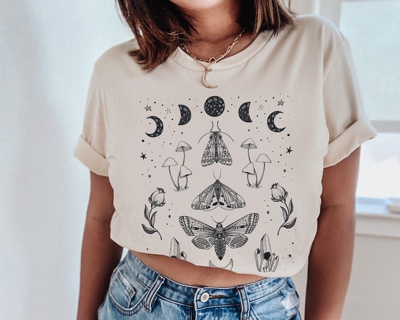 May include: A cream-colored t-shirt with a black and white graphic print of moths, mushrooms, crystals, and crescent moons. The graphic is centered on the shirt.